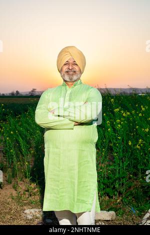 Portrait eines glücklichen Senior-landwirts von punjabi sikh, der im Kreuz mit Turban und Kurta steht und vor die Kamera auf dem Landwirtschaftsfeld schaut. Stockfoto