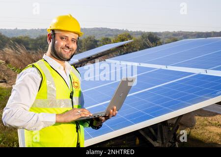 Porträt eines jungen indischen Technikers mit gelbem Schutzhelm, der den Laptop neben Solarpaneelen hält. Installation von Solarsystemen für Industriearbeiter, Stockfoto