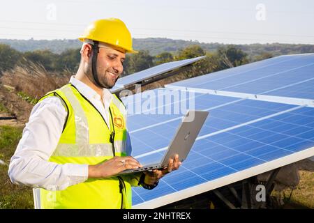 Porträt eines jungen indischen Technikers mit gelbem Schutzhelm, der den Laptop neben Solarpaneelen hält. Installation von Solarsystemen für Industriearbeiter, Stockfoto