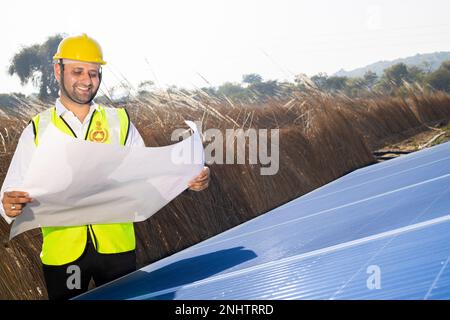 Ein junger indischer Techniker mit gelbem Schutzhelm steht mit Kartenpapier und plant die Installation von Solarpaneelen. Solaranlage für Industriearbeiter Stockfoto