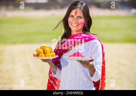 Eine glückliche indische Frau mit Platten voller Laddu- und Pulverfarbe oder Gulal, die das holi Festival im Park feiert. Stockfoto