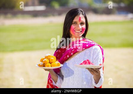 Eine glückliche indische Frau mit Platten voller Laddu- und Pulverfarbe oder Gulal zur Feier des holi Festivals im Park. Stockfoto