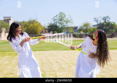 Zwei glückliche junge indische Freundinnen, die ein weißes Kurta-Outfit tragen und mit Pichkari spielen, um das holi-Festival im Park zu feiern. Stockfoto