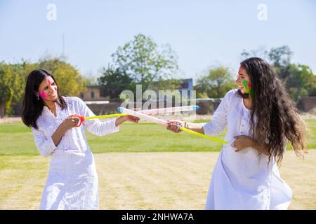 Zwei glückliche junge indische Mädchen, die ein weißes Kurta-Outfit tragen und mit Pichkari spielen, um das holi-Festival im Park zu feiern. Stockfoto
