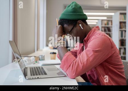 Ein müder afrikanischer Student sitzt vor einem Laptop in der Bibliothek und fühlt sich unmotiviert zu lernen Stockfoto