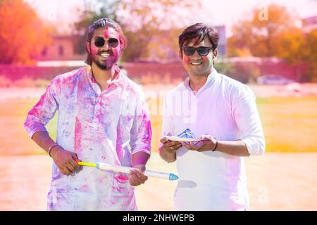 Portrait von glücklichen jungen indischen Männern, die einen weißen Kurta-Halteteller voller Pulverfarbe tragen und Pichkari, die das holi-Festival im Park Outdoor feiern, Face Stockfoto