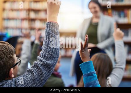 Sie haben eifrige junge Köpfe. Eine Gruppe von Grundschulkindern, die ihre Hand vor ihrem Lehrer in der Bibliothek heben. Stockfoto