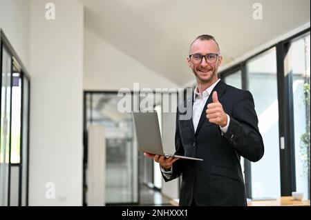 Selbstbewusster und erfolgreicher weißer Geschäftsmann oder männlicher Executive Manager im formellen Business-Anzug, der Daumen nach oben zeigt, ein Notebook in der Hand hält, lächelt und schaut Stockfoto