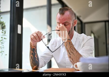 Müder und überarbeiteter weißer Geschäftsmann oder männlicher Büroangestellter sitzt an seinem Schreibtisch, nimmt seine Brille ab, massiert seine Nase und leidet an Aug Stockfoto