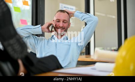 Glücklicher und entspannter männlicher Kaukasier, der seine Beine auf den Tisch und die Hand hinter dem Kopf legte, während er mit dem Kunden im Büro telefonierte. Stockfoto