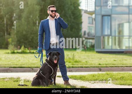 Ein Geschäftsmann, der mit dem Handy telefoniert und seinen Hund an der Leine hält Stockfoto