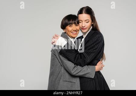 Asiatische Frau mittleren Alters in stilvollen Kleidern, die brünette Tochter umarmt, während sie isoliert auf Grau in die Kamera schaut Stockfoto