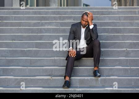 Ein trauriger afroamerikanischer Geschäftsmann, der auf der Treppe vor dem Bürogebäude sitzt, ein Mann in einem Geschäftsanzug, der bankrott gefeuert wurde, der Boss hat Geld verloren. Stockfoto