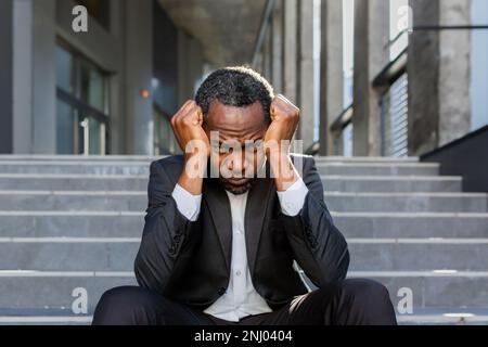 Ein trauriger afroamerikanischer Geschäftsmann, der auf der Treppe vor dem Bürogebäude sitzt, ein Mann in einem Geschäftsanzug, der bankrott gefeuert wurde, der Boss hat Geld verloren. Stockfoto