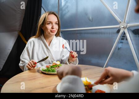 Ein Mädchen im weißen Bademantel frühstückt am Tisch neben dem Fenster. Stockfoto