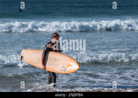 Ein männlicher Surfer, der mit seinem Surfbrett auf dem Fistral in Newquay in Cornwall in England im Vereinigten Königreich aus dem Meer spaziert. Stockfoto