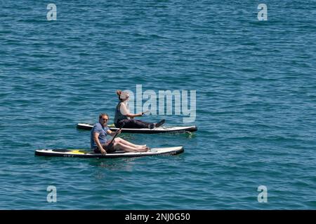 Zwei Urlauber sitzen auf Stand Up Paddleboards in Newquay Bay in Cornwall in England im Vereinigten Königreich. Stockfoto