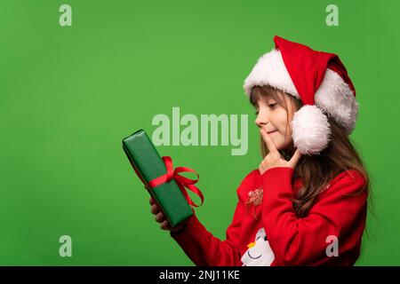 Ein kleines Mädchen mit Weihnachtsmannmütze hält ein Weihnachtsgeschenk in der Hand und versucht, den Inhalt der Schachtel zu erraten. Isoliert auf grünem Hintergrund. Hochwertiges Foto Stockfoto