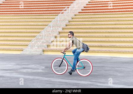 Ein junger Mann trägt eine Sonnenbrille und eine Schultertasche, während er mit dem Fixie Bike fährt. Stockfoto