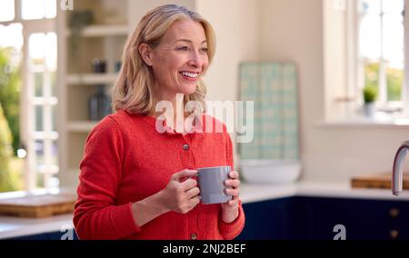 Lächelnde, Reife Frau, Die In Der Küche Steht Und Sich Mit Einer Tasse Kaffee Entspannt Stockfoto