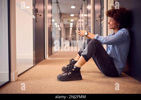 Lächelnde Junge Geschäftsfrau, Die Auf Der Etage Im Korridor Des Modernen Büros Mit Telefon Sitzt Stockfoto