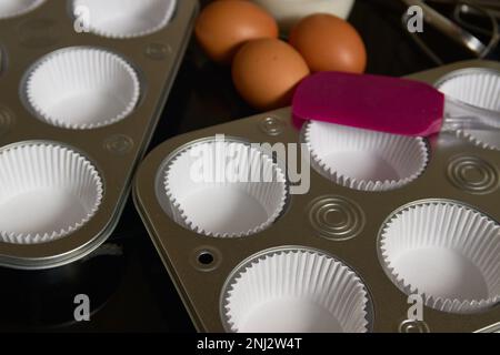 Nahaufnahme einiger Tabletts, die für die Zubereitung von Muffins vorbereitet wurden, zusammen mit braunen Eiern und ein paar Küchenzunge Stockfoto
