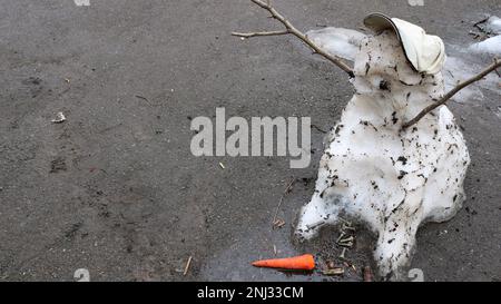 Ein geschmolzener Schneemann in einer Mütze auf grauem Asphalt mit heruntergefallenen Karotten und Metallbolzen als Symbol für den Wechsel der Jahreszeiten oder traurige Emotionen Stockfoto