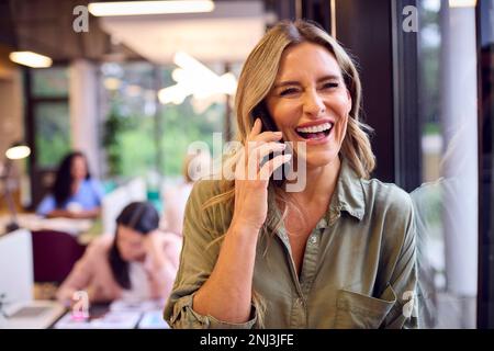 Ältere Geschäftsfrau, Die In Open-Plan-Büros An Der Partition Steht Und Einen Anruf Auf Dem Mobiltelefon Tätigt Stockfoto