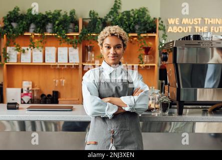 Porträt eines wunderschönen Baristas in einer Schürze. Lächelnder Coffee Shop-Besitzer mit gekreuzten Armen, der sich an der Theke lehnt. Stockfoto