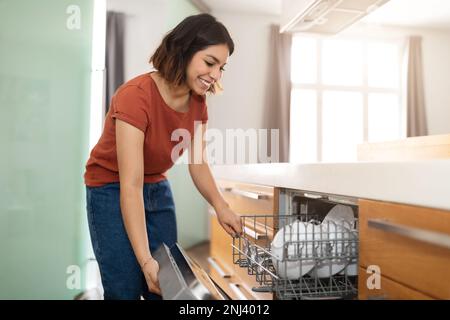 Konzept Der Allgemeinen Ordnung Und Sauberkeit. Junge Lächelnde Araberin, Die Die Spülmaschine In Der Küche Öffnet Stockfoto