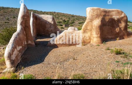 Gekrümmte, verwitterte Lehmwände umschließen einen raumgroßen kiesigen Bereich, mit buschigen Hügeln im Hintergrund, am Fort Bowie National Historic Site, Arizona. Stockfoto