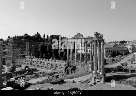 Eine Graustufenaufnahme einer historischen Szene mit einem Uhrenturm auf einer Kirche, umgeben von antiken Ruinen des Forum Romanum Stockfoto