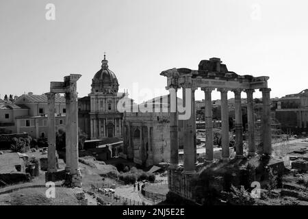 Eine Graustufenaufnahme einer historischen Szene mit einem Uhrenturm auf einer Kirche, umgeben von antiken Ruinen des Forum Romanum Stockfoto