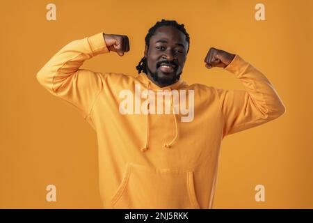 Ein lächelnder Afrikaner in einem gelben Hoodie zeigt seine körperliche Stärke vor gelbem Hintergrund Stockfoto