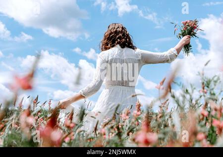 Rückansicht einer Frau in langem weißen Kleid mit einem Wildblumenstrauß auf einem rosafarbenen Blumenfeld Stockfoto