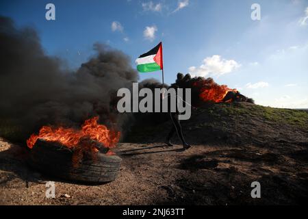 Gaza-Stadt. Palästina. 22. Februar 2023. Ein Demonstrant hält die palästinensische Flagge vor brennenden Reifen während einer Demonstration entlang der Grenze zwischen dem Gazastreifen und Israel, in Solidarität mit der Stadt Nablus gegen die israelischen Militäroperationen, bei denen mindestens zehn Palästinenser ums Leben kamen. Stockfoto