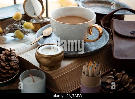 Erfrischend am Morgen mit heißem Kaffee in Keramiktasse und Messing-Kaffeelöffel serviert mit frischer Milch auf einem Arbeitstisch mit altem Buch, Messingkompass, Duftdose Stockfoto