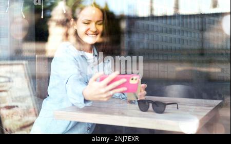 Frau sitzt im Café und macht ein Selfie mit ihrem Smartphone, reflektiert die Straße auf einem Glas Café. Stockfoto