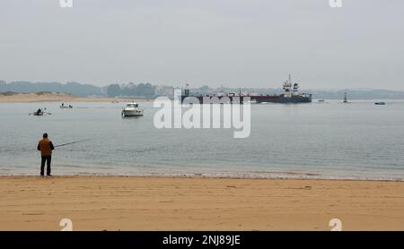 Ein Mann, der vom Strand aus angeln kann, als Cemvale Cement Carrier Frachtschiff, das den Hafen von Santander an einem ruhigen, nebligen Wintermorgen Santander Cantabria Spanien verlässt Stockfoto