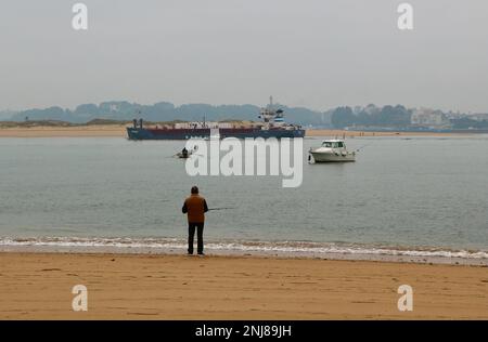 Ein Mann, der vom Strand aus angeln kann, als Cemvale Cement Carrier Frachtschiff, das den Hafen von Santander an einem ruhigen, nebligen Wintermorgen Santander Cantabria Spanien verlässt Stockfoto