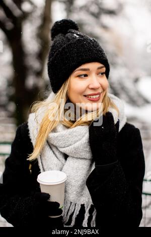 Eine junge Frau steht in einem Winterwunderland, trägt warme Kleidung und trinkt heißen Kaffee zum Mitnehmen Stockfoto