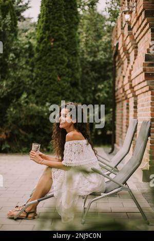 Hübsche junge Frau mit lockigem Haar entspannt sich auf einem Liegestuhl und genießt ein Glas frische Limonade Stockfoto