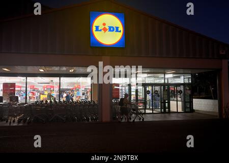 Vor einem Lidl-Einzelhandelsgeschäft bei Nacht mit beleuchtetem Innenraum und Silhouetten von Einkaufswagen und Kunden draußen Stockfoto