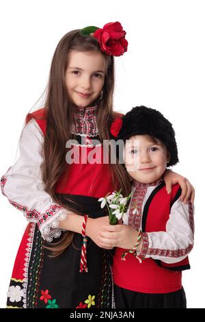 Bulgarische Kinder Jungen und Mädchen in traditionellen Folklore-Kostümen mit Frühlingsblumen, Schneetropfen und handgefertigten Wollarmband Martenitsa Symbol von Baba Marta Stockfoto