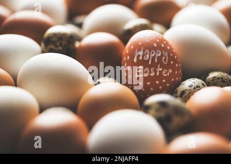 Happy easter schrieb weißes Hühnerei auf einen Haufen Eier. Ein Stapel brauner und weißer Hühnereier mit Wachteleiern Stockfoto