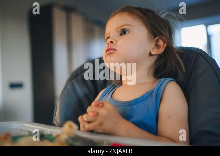 Süßes niedliches Baby Mädchen, das in einem Hochstuhl sitzt und in der Küche zu Hause unabhängig isst. Authentische Momente. Gesundes Essen. Gesunde Ernährung. Stockfoto