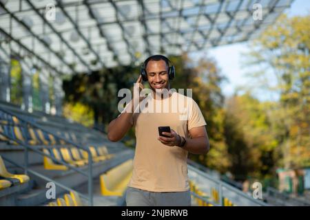 Ein junger afroamerikanischer Sportler, Sportler und Läufer mit Kopfhörern steht im Stadion zwischen den Tribünen. Hält das Telefon in seinen Händen, hört Musik. Er schloss die Augen und ruhte sich aus. Stockfoto