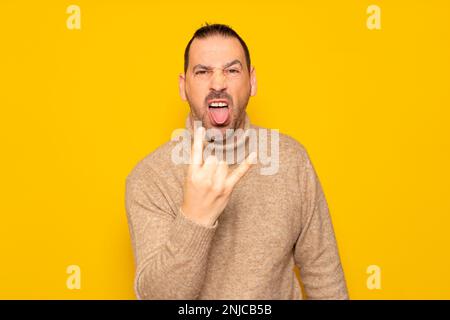 bärtiger lateinamerikanischer Mann mit Rollkragenpullover, isoliert auf gelbem Hintergrund und Geste mit Fingern und herausragender Zunge. Konzept von Stockfoto