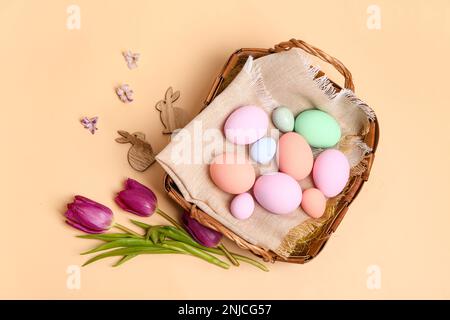 Korb aus Korb mit bemalten Ostereiern, Hasen und Tulpenblüten auf beigefarbenem Hintergrund Stockfoto