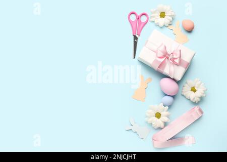 Geschenk mit Schere, Ostereiern, Papierkaninchen und Blumen auf blauem Hintergrund Stockfoto